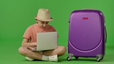 Şapkalı genç çocuk mor bavulun yanında oturuyor ve dizüstü bilgisayarda yazıyor. Krom anahtar yeşil ekran yeşil ekran stüdyo arka plan modelinde izole edilmiş. Yavaş çekim