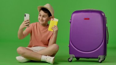 Şapkalı genç, mor bavulun yanında oturuyor. Pasaportunu ve uçak biletlerini gösteriyor. Telefonda selfie çekiyor. Kromakey yeşil ekranda izole edilmiş, yeşil ekran, stüdyo arka planı, maket. Yavaş çekim