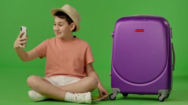 Mor bavulun yanında oturan şapkalı genç çocuk akıllı telefondan selfie çekiyor. Krom anahtar yeşil ekran yeşil ekran stüdyo arka plan modelinde izole edilmiş. Yavaş çekim