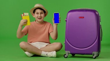 Şapkalı genç bir çocuk mor bavulun yanında oturuyor, elinde pasaportu ve akıllı telefonu var. Krom anahtar yeşil ekran yeşil ekran stüdyo arka plan modelinde izole edilmiş. Yavaş çekim