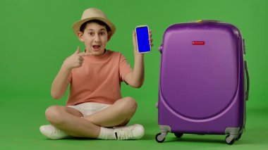 Mor bavulun yanında oturan şapkalı genç çocuk mavi ekranlı akıllı telefonu işaret ediyor. Krom anahtar yeşil ekran yeşil ekran stüdyo arka plan modelinde izole edilmiş. Yavaş çekim