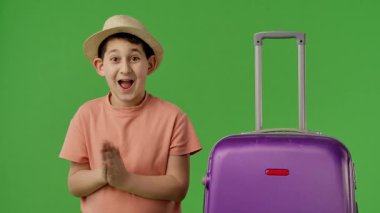 Şapkalı bir çocuk, parlak yeşil bir arka planda mor bir bavulun yanında alkışlar ve sevinir. Yavaş çekim