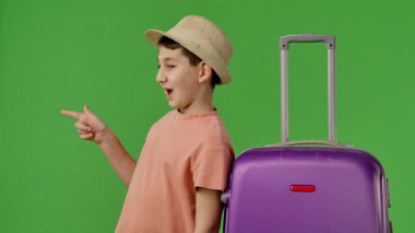 Şapkalı genç bir çocuk mor bavulun yanında duruyor ve parmağıyla yeri işaret ediyor. Kopya uzayı. Krom anahtar yeşil ekran yeşil ekran stüdyo arka plan modelinde izole edilmiş. Yavaş çekim