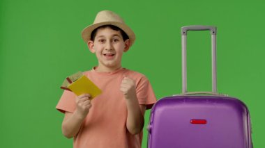 Mor bavulun yanında duran şapkalı genç çocuk pasaport ve uçak biletlerini gösteriyor. Krom anahtar yeşil ekran yeşil ekran stüdyo arka plan modelinde izole edilmiş. Yavaş çekim