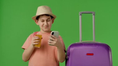 Şapkalı neşeli bir çocuk bavulunun yanında meyve suyu ve akıllı telefon tutuyor, tatili için heyecanlı. Yavaş çekim