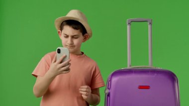 Mor bavulun yanında duran şapkalı genç çocuk akıllı telefondan selfie çekiyor. Krom anahtar yeşil ekran yeşil ekran stüdyo arka plan modelinde izole edilmiş. Yavaş çekim