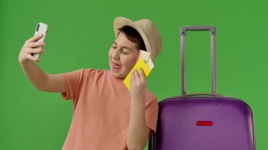 Şapkalı genç, mor bavulun yanında duruyor. Pasaportunu ve uçak biletlerini gösteriyor. Telefonda selfie çekiyor. Kromakey yeşil ekranda izole edilmiş, yeşil ekran, stüdyo arka planı, maket. Yavaş çekim