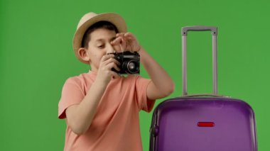 Şapka takan bir çocuk bavulun yanında fotoğraf makinesi tutuyor ve heyecan verici bir seyahat macerasına hazır. Yavaş çekim