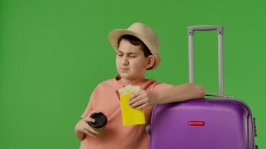 Şapkalı genç bir çocuk, fincandan su içiyor, bavula yaslanıyor, pasaportuyla seyahat etmeye hazır. Yavaş çekim