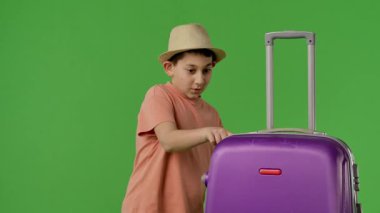 Hasır şapka takan genç bir çocuk büyük mor bir bavulun üzerinde yeşil bir zemin üzerinde dinleniyor, bitkin görünüyor. Yavaş çekim