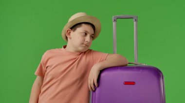 Hasır şapka takan genç bir çocuk büyük mor bir bavulun üzerinde yeşil bir zemin üzerinde dinleniyor, bitkin görünüyor. Yavaş çekim