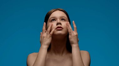 Çıplak omuzlu bir kadın, yukarı bakarken yüzüne nazikçe masaj yapar. Düz mavi bir arka plana bakar..