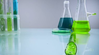 Parlak bir laboratuvar masasına yeşil sıvı sızdıran parçalanmış bir test tüpünü kapat. Arka planda renkli çözeltilere ve bitki örneklerine sahip diğer test tüpleri ve mataralar görülebilir..