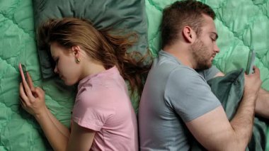 Bir çift sırt sırta yatar, her ikisi de kendi akıllı telefonlarına gömülüp dijital bağlantısızlığı vurgular. Yavaş çekim