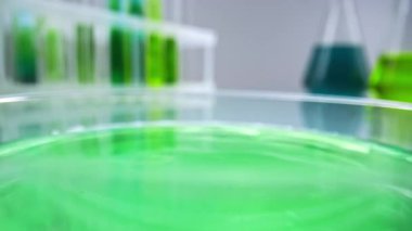 Bir damla yeşil sıvı bir laboratuvar yüzeyindeki petri kabında sıçrama oluşturur. Arka planda bulanık test tüpleri ve renkli sıvılarla dolu mataralar bilimsel araştırmaları temsil ediyor..
