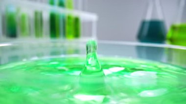 Bir damla yeşil sıvı bir laboratuvar yüzeyindeki petri kabında sıçrama oluşturur. Arka planda bulanık test tüpleri ve renkli sıvılarla dolu mataralar bilimsel araştırmaları temsil ediyor..