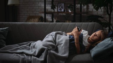 Bir kadın rahat bir şekilde battaniyenin altındaki kanepeye uzanır, rahat bir odada akıllı telefonunu karıştırmaya odaklanır..
