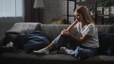 Kanepede tek başına oturan bir kadın düşünceli ve düşünceli görünüyor. Oda loş bir şekilde aydınlatılmış, yansıtıcı bir atmosfer yaratıyor..