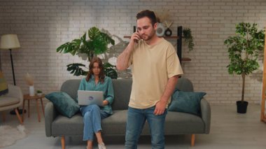 Bir erkek telefonda konuşurken, bir kadın dizüstü bilgisayarı üzerinde çalışıyor. Parlak, modern bir oturma odasında..