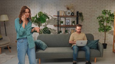 Modern bir oturma odasında telefonla konuşan bir kadın, bir erkeğin dizüstü bilgisayarla çalışması gibi hayal kırıklığına uğramış görünür..