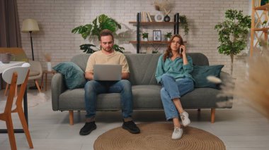 Bir adam ve bir kadın gri bir kanepede oturuyorlar. Kadın telefonda konuşurken adam dizüstü bilgisayar kullanıyor..