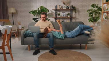 Bir çift modern bir oturma odasındaki kanepede dinleniyor, biri dizüstü bilgisayarda çalışıyor, diğeri de huzur içinde kitap okuyor..