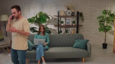 Bir erkek telefonda konuşurken, bir kadın dizüstü bilgisayarı üzerinde çalışıyor. Parlak, modern bir oturma odasında..