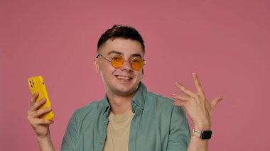 Şık güneş gözlüklü gülümseyen genç bir adam sarı bir akıllı telefon tutuyor, canlı pembe bir arkaplanda poz veriyor, kendine güveni ve tarzı var..