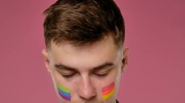 Pembe arka planda yüzü gökkuşağı gibi olan genç bir adam başını eğip kameraya bakıyor ve LGBT gururunu ve kimliğini simgeliyor..