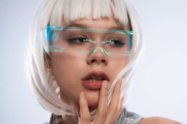 Beyaz kısa saçlı, neon ışıklı gözlüklü, yüzüne dokunan gelecekten gelen bir kadın. Gümüş metalik kıyafetler giyiyor, modern bilim kurgu modasını ve gelişmiş gerçeklik teknolojisini sergiliyor..