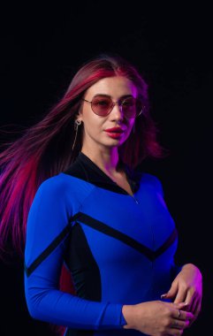 Pembe saçlı, parlak mavi elbiseli, yuvarlak güneş gözlüklü, esmer arka planda poz veren modern bir kadın. Fütürist moda ve siber punk estetiği.