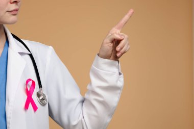Beyaz önlüklü, steteskoplu ve pembe kurdeleli bir doktor tarafsız bir arkaplanı işaret ediyor. Göğüs kanseri farkındalık günü konsepti.