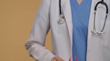 Bej arka planda beyaz önlüklü bir doktor, farkındalığın sembolü olarak kameraya pembe kurdeleli bir el uzatıyor. Göğüs kanseri farkındalık günü, kadın sağlığı ve toplum konsepti.