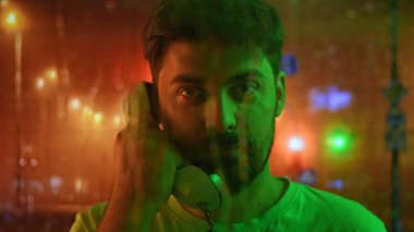 Telefon kulübesinde konuşan sakallı bir adam. Ön plandaki bardaktan damlayan yağmur damlaları, arka plandaki akşam şehrinin bulanık ışıkları. Retro kavramı.