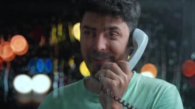 Sakallı bir adam telefon kulübesinde mutlu bir şekilde konuşuyor. Ön planda bardaktan damlayan yağmur damlaları, arka planda akşam şehrinin bulanık ışıkları. Retro kavramı.