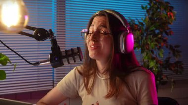 Modern bir stüdyoda podcast yaparken kulaklık takan ve mikrofona konuşan genç bir kadın. Yavaş çekim