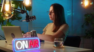 Bir kadın şık bir stüdyoda podcast yapıyor. Mikrofon, dizüstü bilgisayar kullanıyor ve kulaklık takıyor. Bir gösteri, sunum ya da video içeriği kaydediliyor. Yavaş çekim