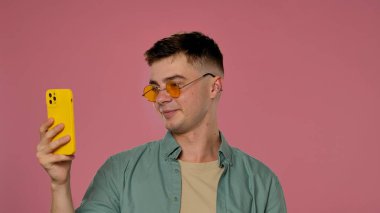 Sarı güneş gözlüğü takan bir adam akıllı telefonuyla pembe arka planda poz vererek şık ve modaya uygun bir stüdyo ortamı yaratıyor..