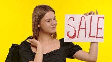 Elinde alışveriş torbaları olan genç bir kadın, parlak sarı bir zemin üzerinde indirim öneren bir satış tabelası tutuyor..