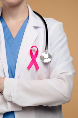 Beyaz önlüklü, steteskop ve pembe kurdeleli, kolları güvenle bağlanmış bir doktor. Göğüs kanseri farkındalık günü konsepti.