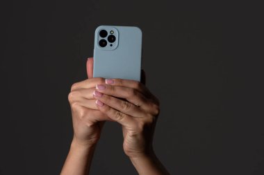 Bir çift el, açık renkli çantalı modern bir akıllı telefonu tutuyor, karanlık bir arkaplanda, anı yakalamaya odaklanıyor..