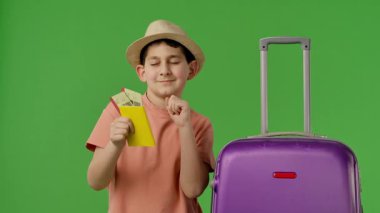 Mor bavulun yanında duran şapkalı genç çocuk pasaport ve uçak biletlerini gösteriyor. Krom anahtar yeşil ekran yeşil ekran stüdyo arka plan modelinde izole edilmiş. Yavaş çekim