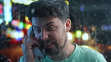 Sakallı bir adam telefon kulübesinde negatif konuşuyor. Ön planda damlayan yağmur damlaları, arka planda şehrin bulanık ışıkları. Retro kavramı.
