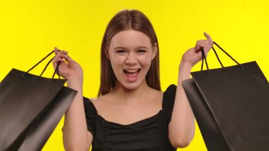 Neşeli genç bir kadın, parlak bir gülümsemeyle iki siyah alışveriş torbasını canlı sarı arka planda tutuyor..