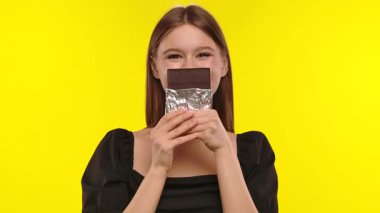 Bir kadın neşeyle folyoya sarılı çikolatayı canlı sarı bir arka fona tutarak zevkini ifade eder..