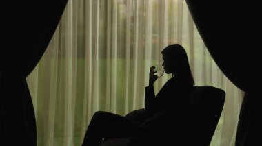 Yumuşak ışıklandırmalı ve perdeli bir pencereye yaslanmış, bardaktan su içen bir kadının sakin silueti rahatlatıcı bir atmosfer yaratıyor. Akıl sağlığı kavramı.
