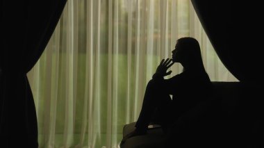 Yumuşak ışıklı perdelerin önünde oturan bir kadının silueti üzgün, stresli ve içe dönük ifade ediyor. Akıl Sağlığı Konsepti.