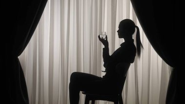 Yumuşak perdelerin arkasına bir bardak su tutarak sakinleştiren bir atmosfer yaratan huzurlu bir kadın silueti. Ruh sağlığı konsepti.