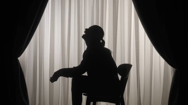 Yumuşak ışıklı perdelerin önünde oturan bir kadının silueti üzgün, stresli ve içe dönük ifade ediyor. Akıl Sağlığı Konsepti.