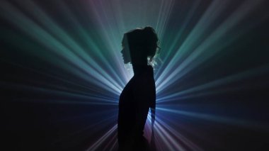 Bir kadının dramatik bir silueti canlı bir gökkuşağı ışık projeksiyonuna karşı duruyor, gizemli, sanatsal bir kompozisyonda vücudunun ana hatlarını belirten parlak bir renk patlaması yaratıyor. Yan görünüm.